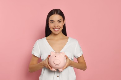 Happy young woman with ceramic piggy bank on pale pink background