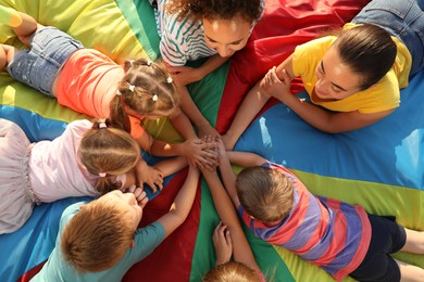 Photo of Group of children with teachers holding hands together on rainbow playground parachute, top view. Summer camp activity