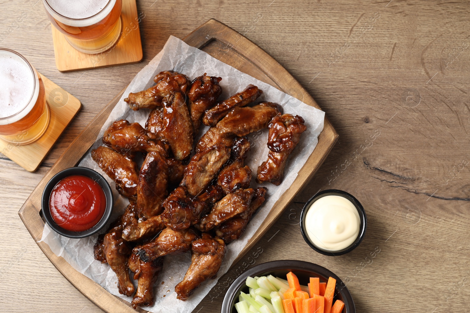 Photo of Tasty chicken wings, glasses of beer, sauces and vegetable sticks on wooden table, flat lay. Delicious snacks