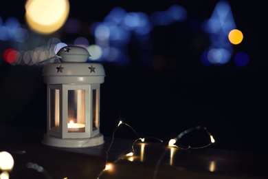Photo of Lantern and Christmas lights on wooden railing outdoors against blurred background, space for text. Winter night