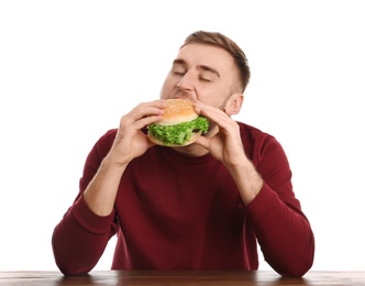 Young man eating tasty burger at table on white background