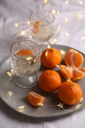 Photo of Delicious ripe tangerines, glasses of sparkling wine and fairy lights on white bedsheet