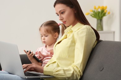 Mother working remotely on laptop while her daughter playing with smartphone at home