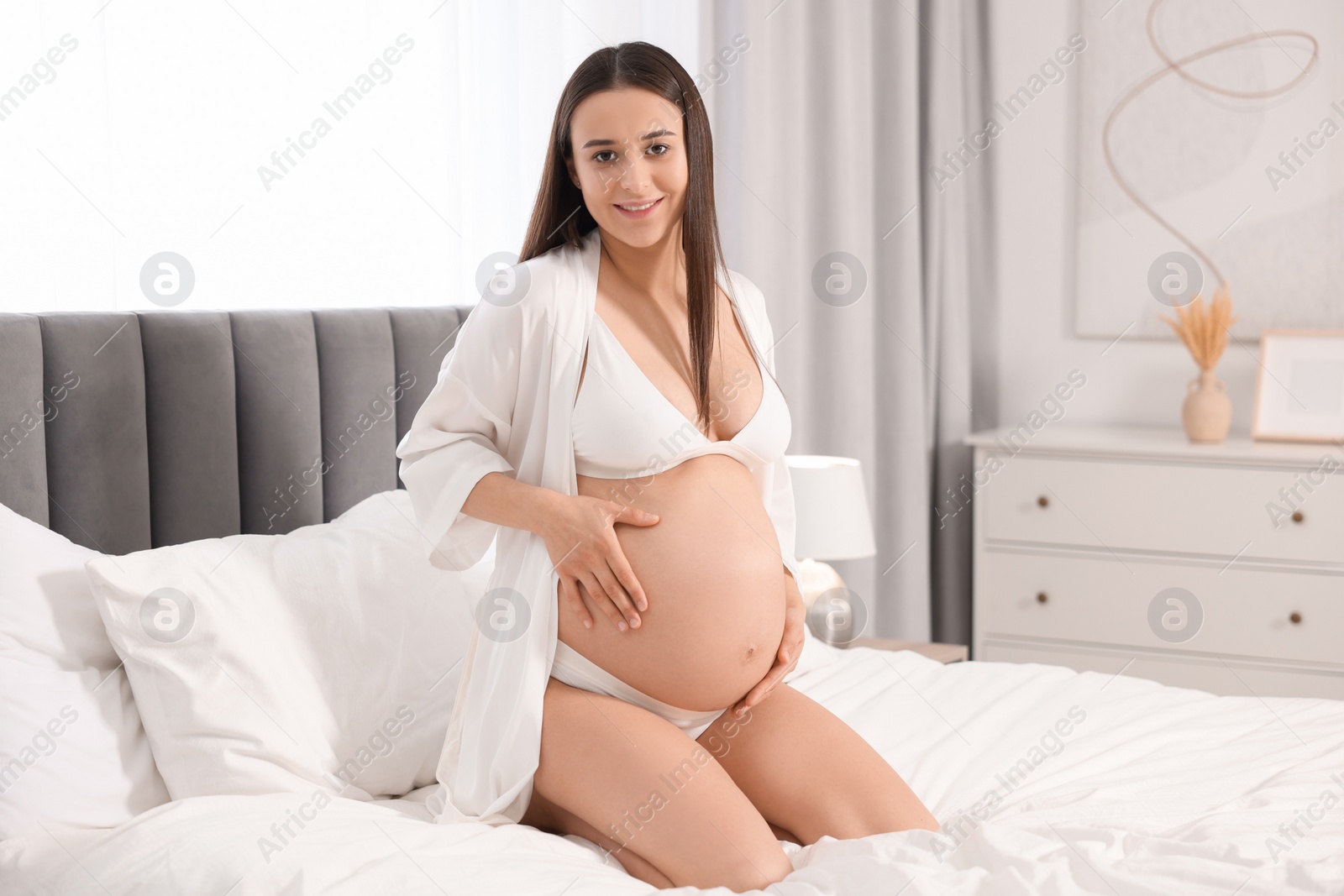 Photo of Beautiful pregnant woman in stylish comfortable underwear and robe on bed at home, space for text