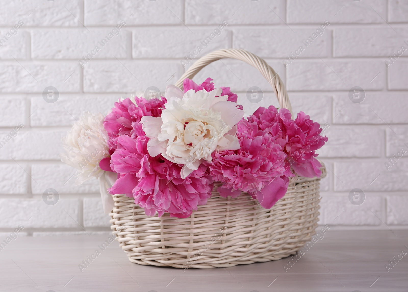 Photo of Beautiful peonies in wicker basket on white table near brick wall