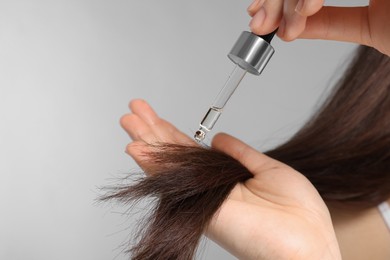 Photo of Woman applying essential oil onto hair on light grey background, closeup