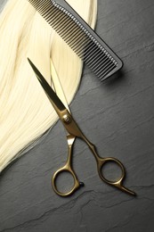Photo of Professional hairdresser scissors and comb with blonde hair strand on dark grey table, flat lay