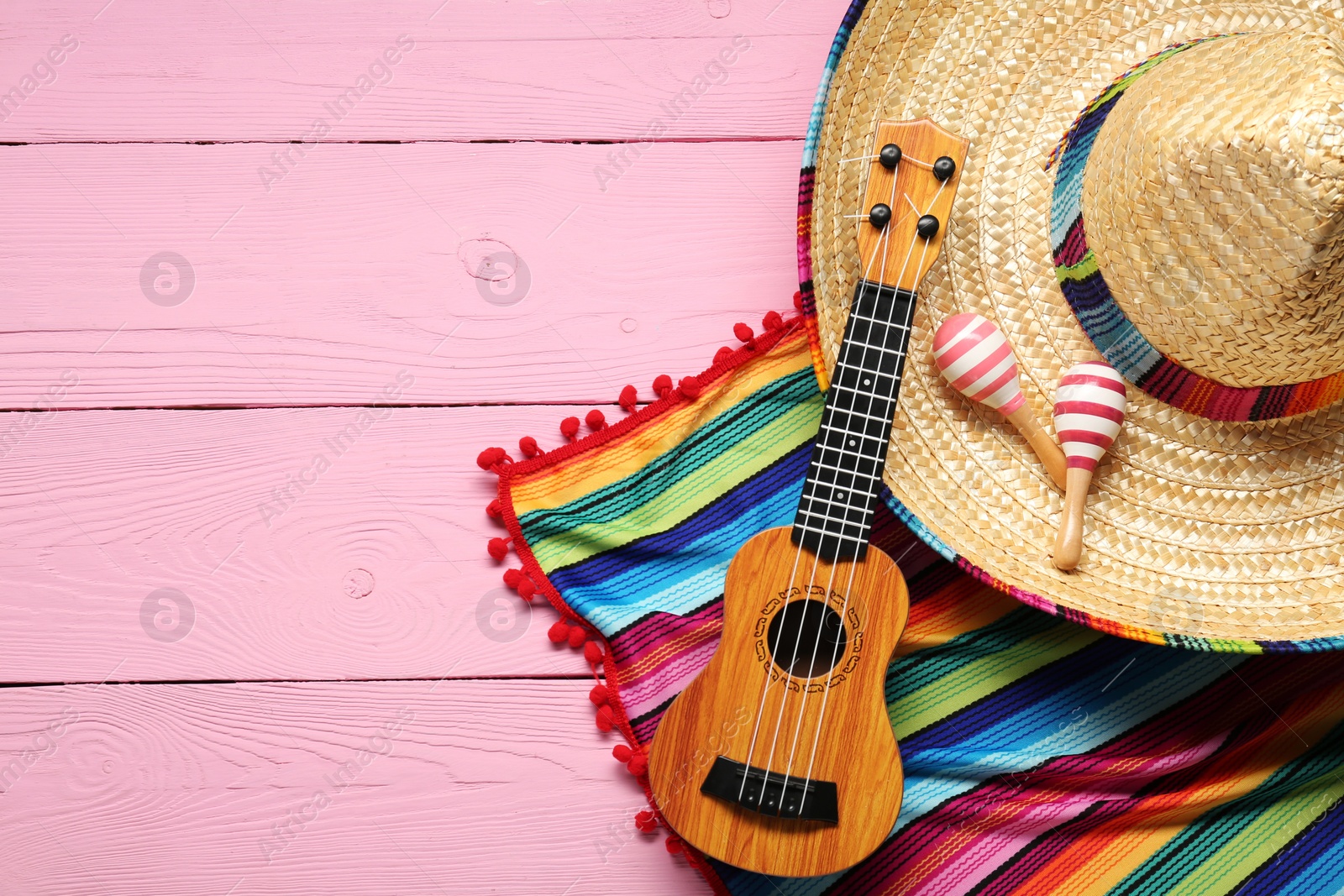 Photo of Mexican sombrero hat, guitar, maracas and colorful poncho on pink wooden background, flat lay. Space for text