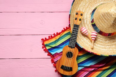Mexican sombrero hat, guitar, maracas and colorful poncho on pink wooden background, flat lay. Space for text