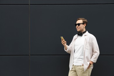 Photo of Handsome young man with headphones and modern smartphone near black wall. Space for text