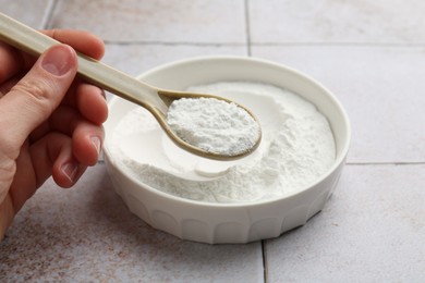 Woman taking baking powder with spoon from bowl at light tiled table, closeup