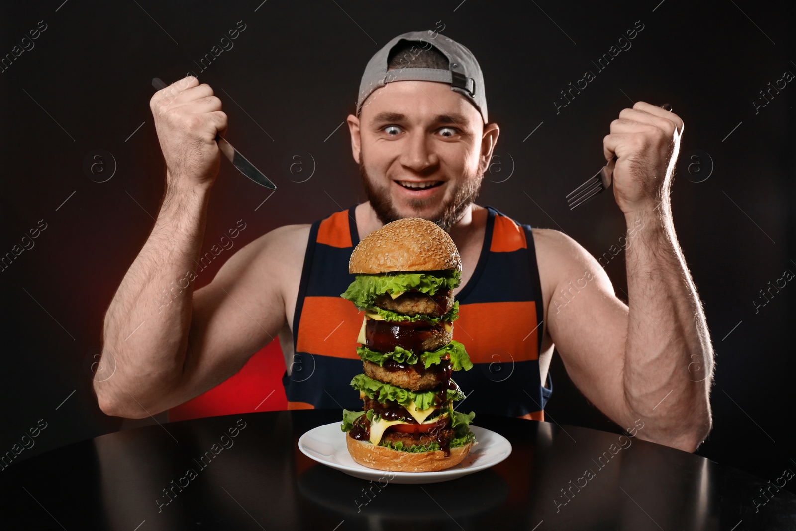 Photo of Young hungry man with cutlery eating huge burger on black background