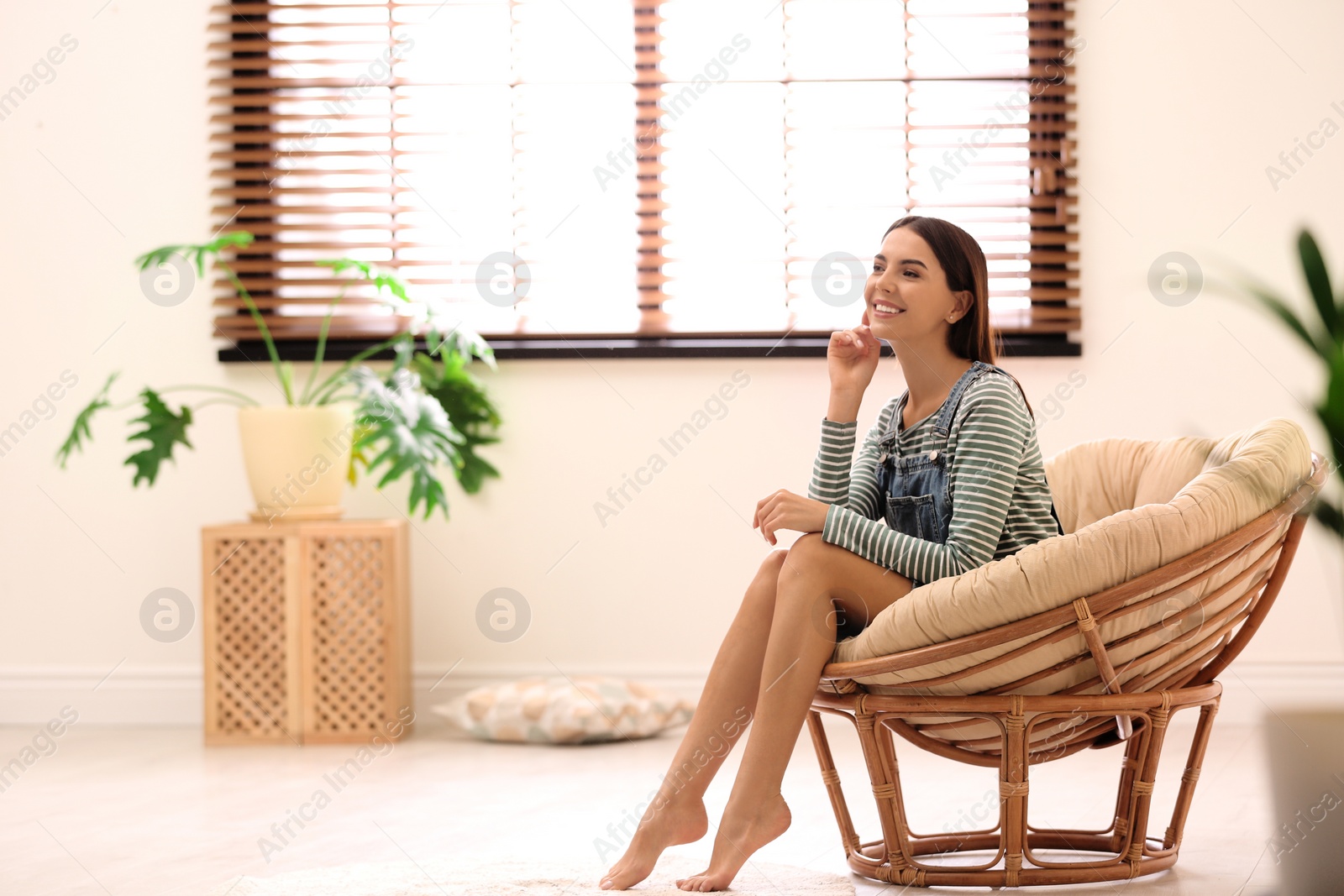 Photo of Young woman relaxing in papasan chair near window with blinds at home. Space for text