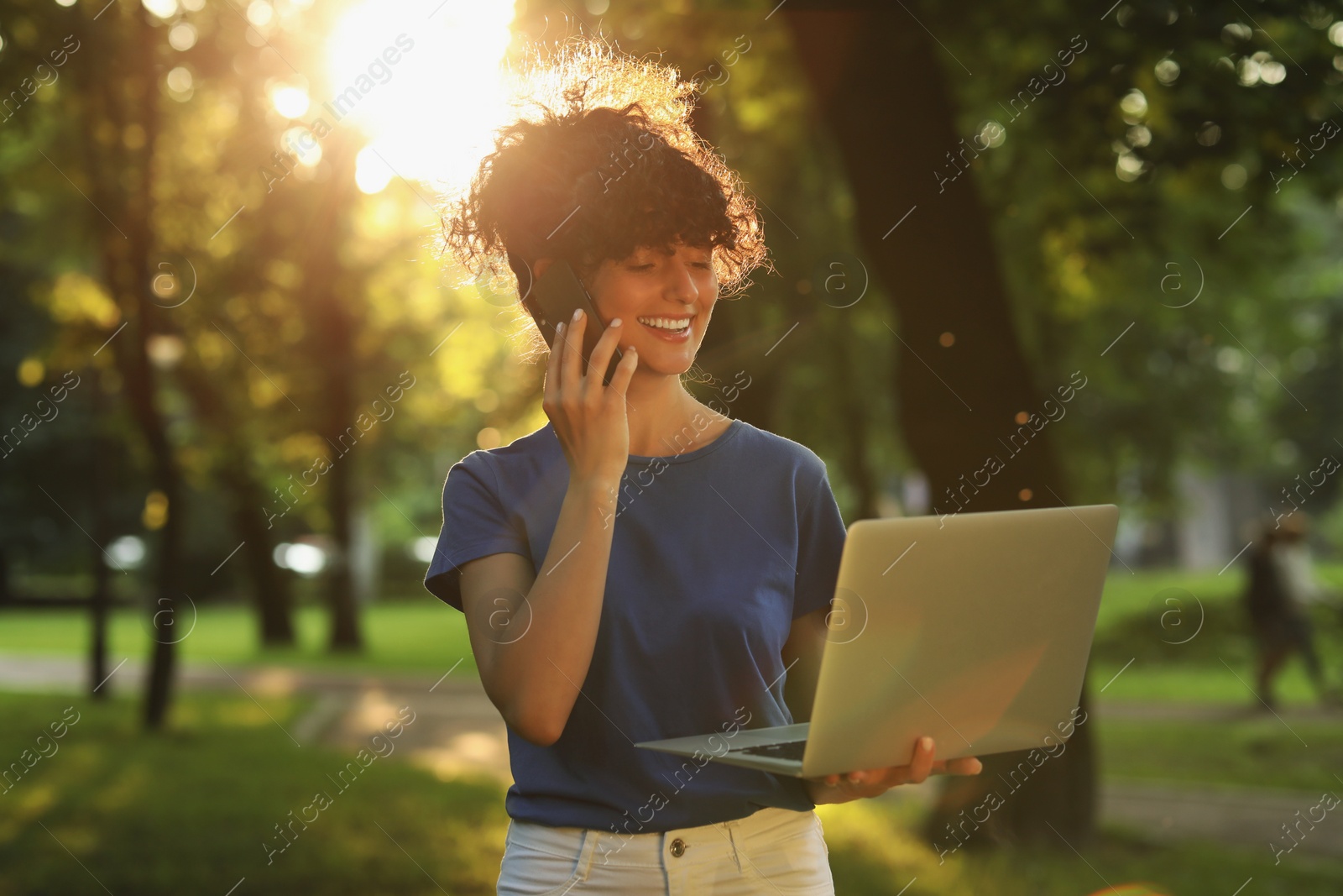 Photo of Happy young woman using modern laptop and talking on smartphone in park