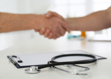 Closeup view of doctor shaking hands with senior patient in office, focus on clipboard and stethoscope