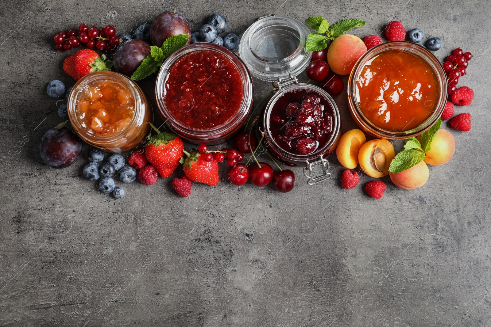 Photo of Jars with different jams and fresh fruits on grey table, flat lay. Space for text