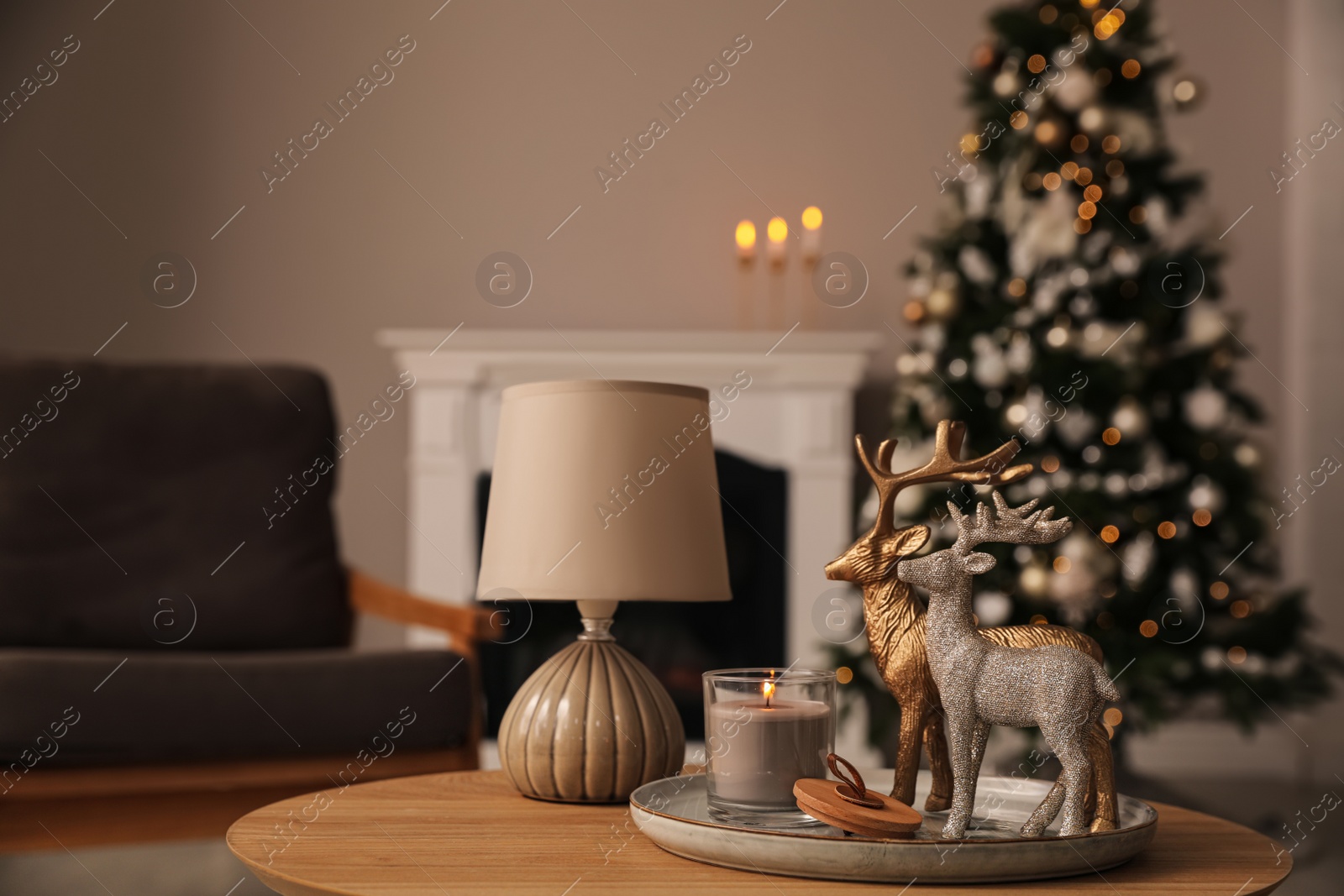 Photo of Deer figures, burning candle with lamp on wooden table and Christmas tree in living room