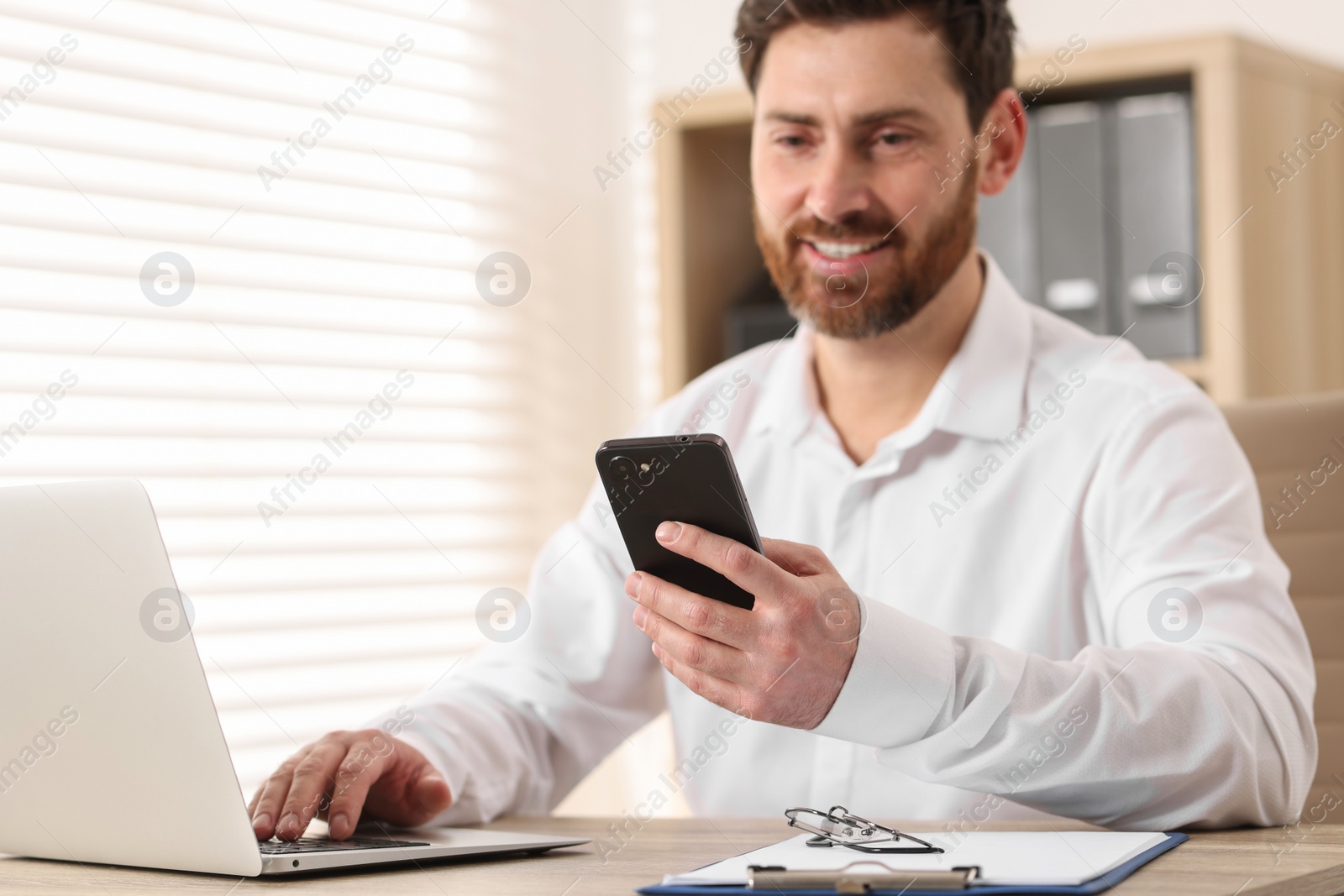 Photo of Smiling man with smartphone using laptop at table in office, selective focus