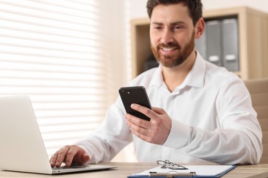 Smiling man with smartphone using laptop at table in office, selective focus