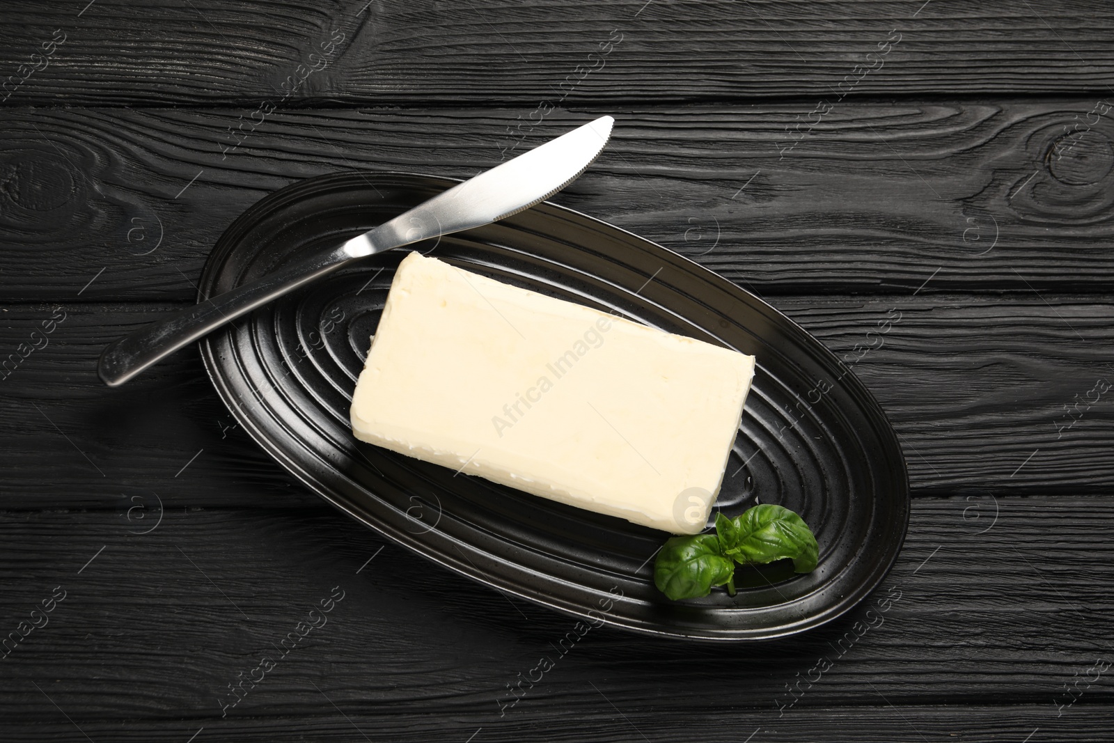 Photo of Block of tasty butter with basil and knife on black wooden table, top view