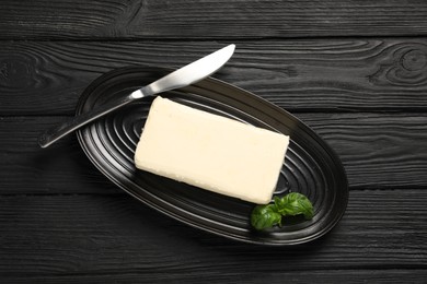 Block of tasty butter with basil and knife on black wooden table, top view