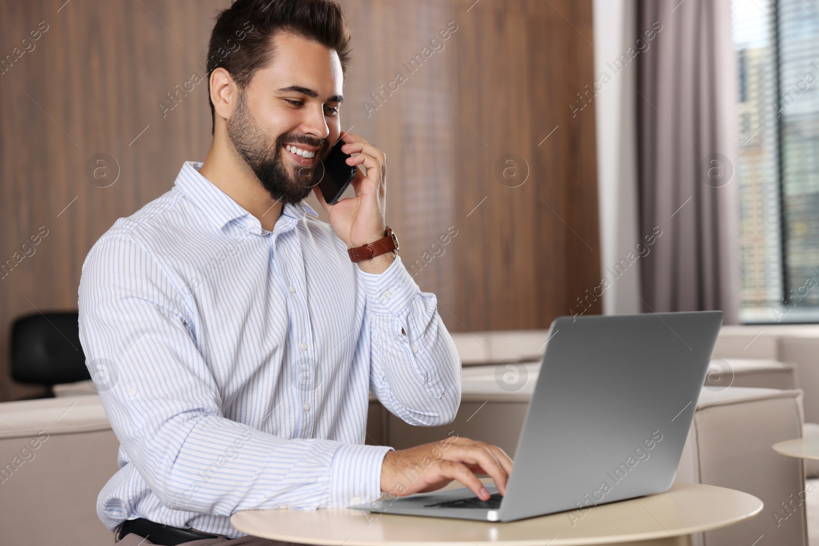 Photo of Happy man using modern laptop while talking on smartphone at table in office