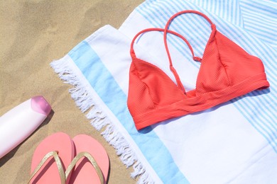 Photo of Striped towel with bikini top, bottle of sunblock and flip flops on sandy beach, flat lay