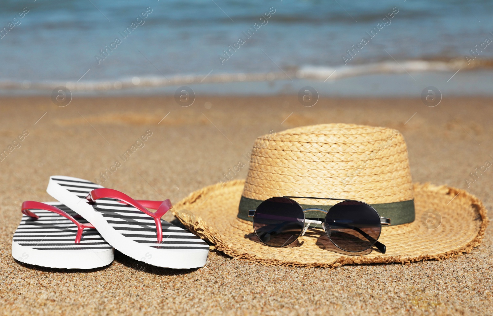 Photo of Striped flip flops, straw hat and sunglasses on sandy beach near sea