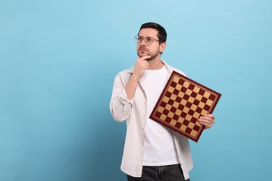 Thoughtful man holding chessboard on light blue background. Space for text
