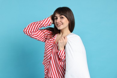 Happy woman wearing pyjama and holding blanket on light blue background