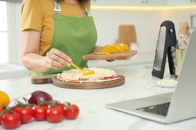 Photo of Woman making pizza while watching online cooking course via laptop in kitchen, closeup