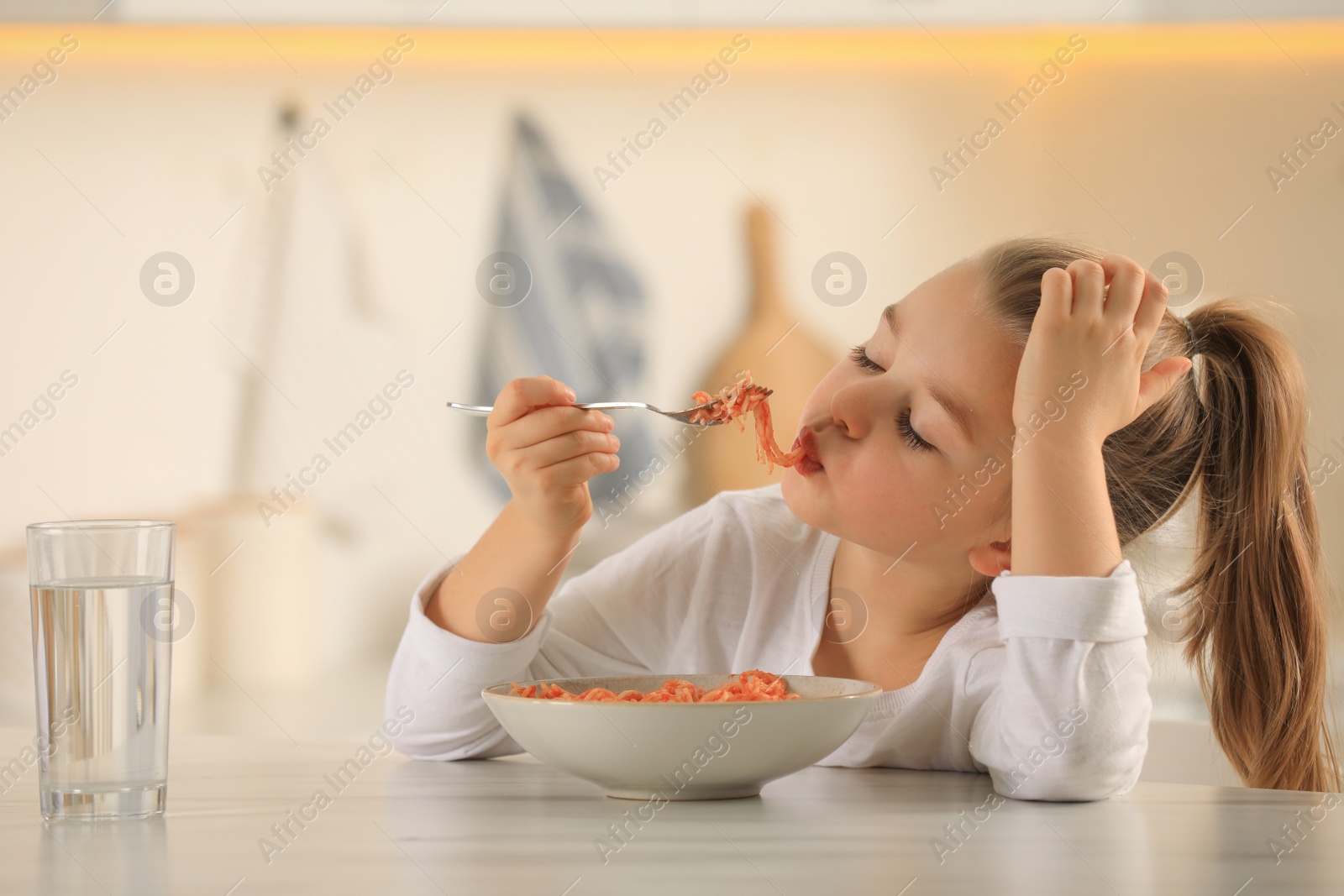 Photo of Cute little girl eating tasty pasta at table in kitchen