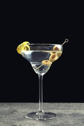 Photo of Martini cocktail with olives and lemon twist on grey table against dark background