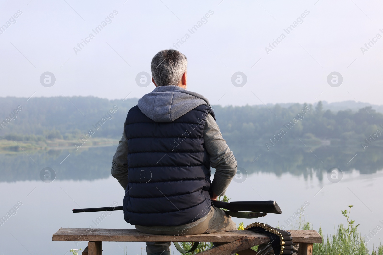 Photo of Man with hunting rifle sitting on wooden bench near lake outdoors, back view