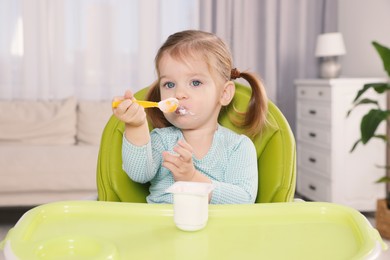 Cute little child eating tasty yogurt from plastic cup with spoon in high chair indoors