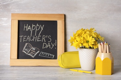 Chalkboard with inscription HAPPY TEACHER'S DAY, stationery and vase of flowers on wooden table against light wall