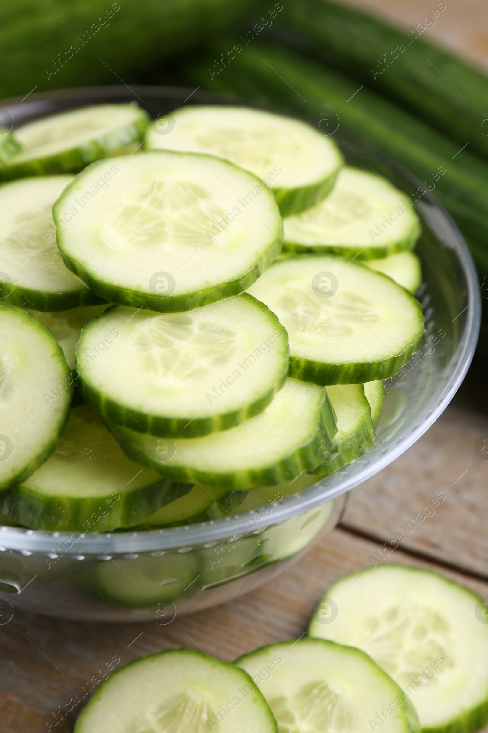 Photo of Cut cucumber in glass bowl and fresh vegetables on wooden table, closeup