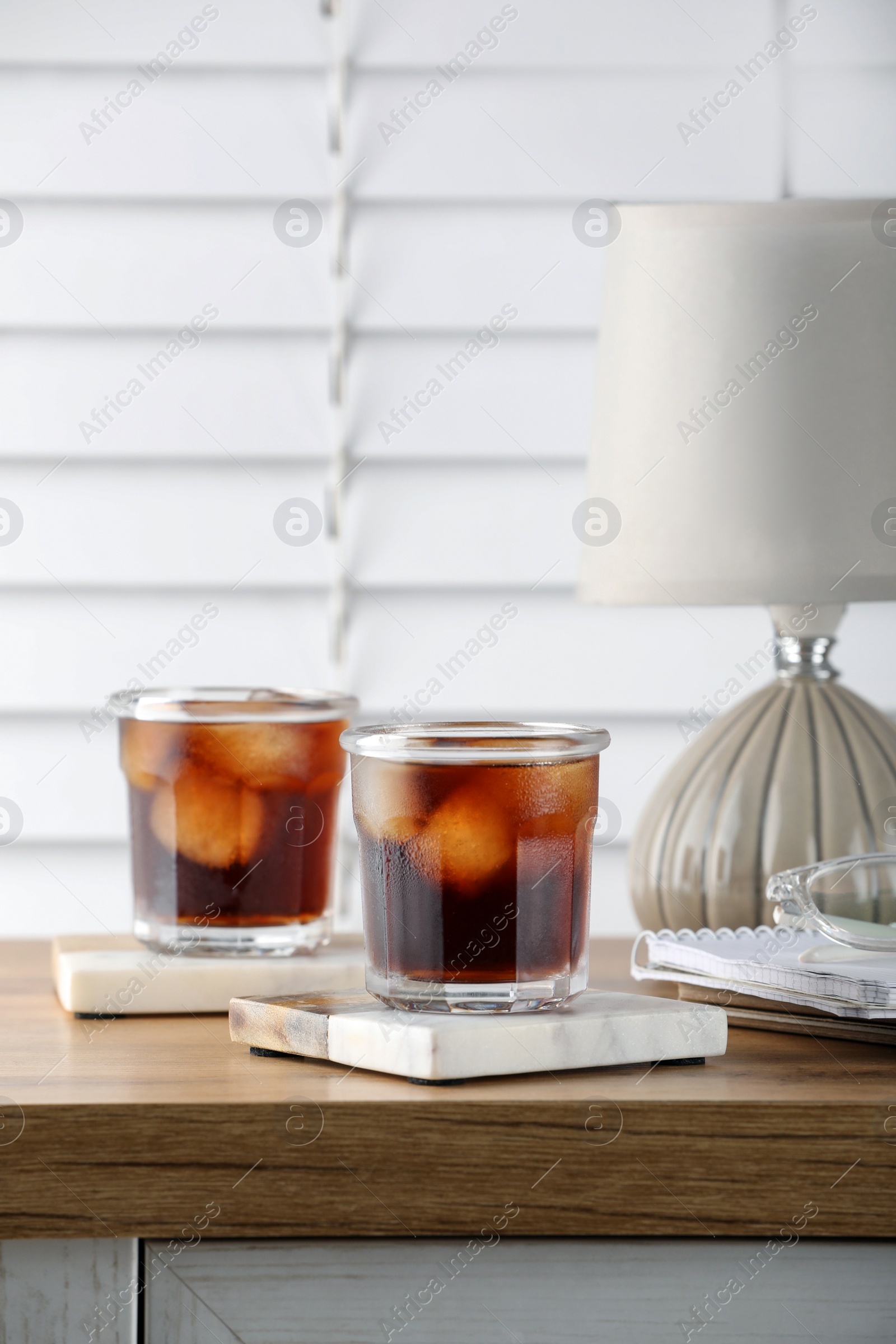 Photo of Glasses of cold drink with stylish cup coasters, lamp and stationery on wooden table in room