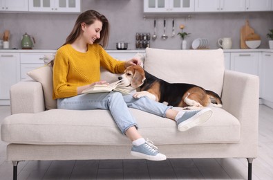Photo of Happy young woman reading book near her cute Beagle dog on couch at home. Lovely pet