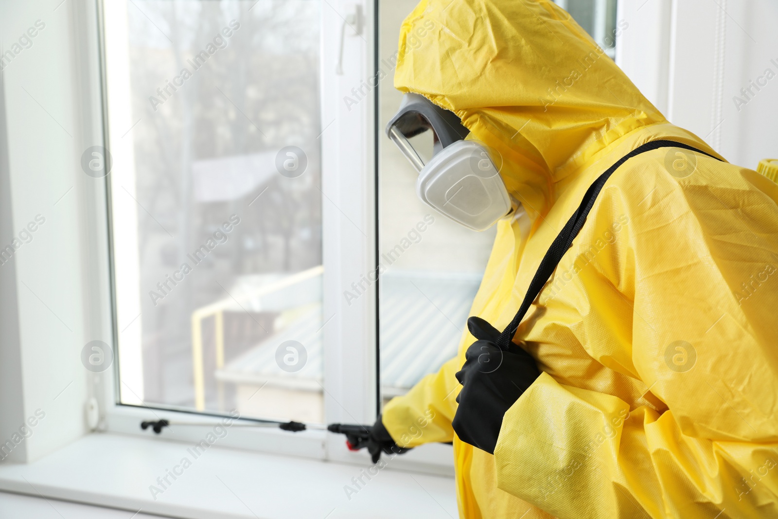 Photo of Pest control worker in protective suit spraying insecticide on window sill indoors