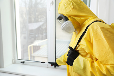 Photo of Pest control worker in protective suit spraying insecticide on window sill indoors