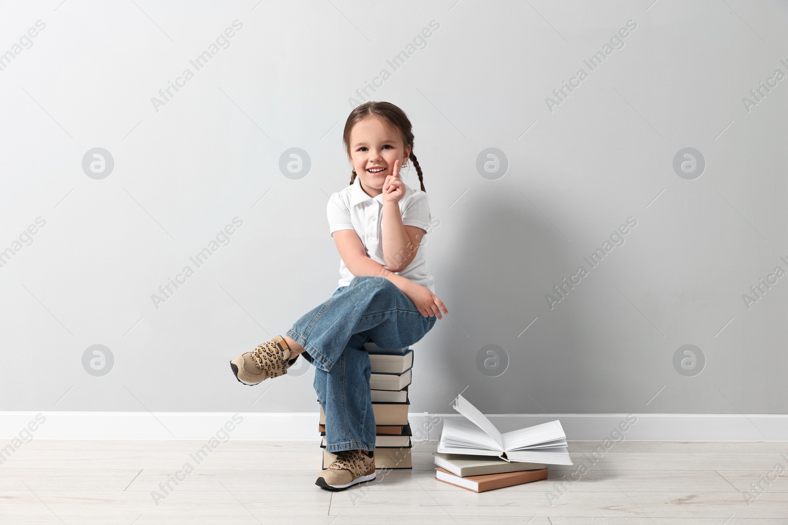 Photo of Cute little girl sitting on stack of books near light grey wall