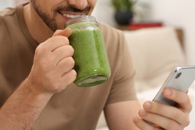 Photo of Happy man holding delicious fresh smoothie and using smartphone indoors, closeup