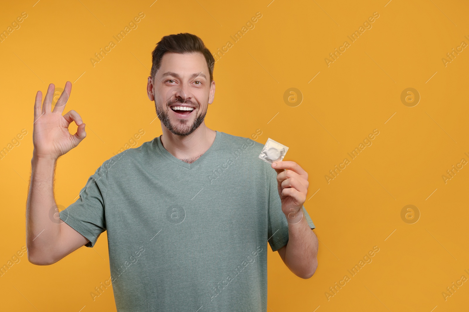 Photo of Happy man with condom showing ok gesture on orange background. Space for text
