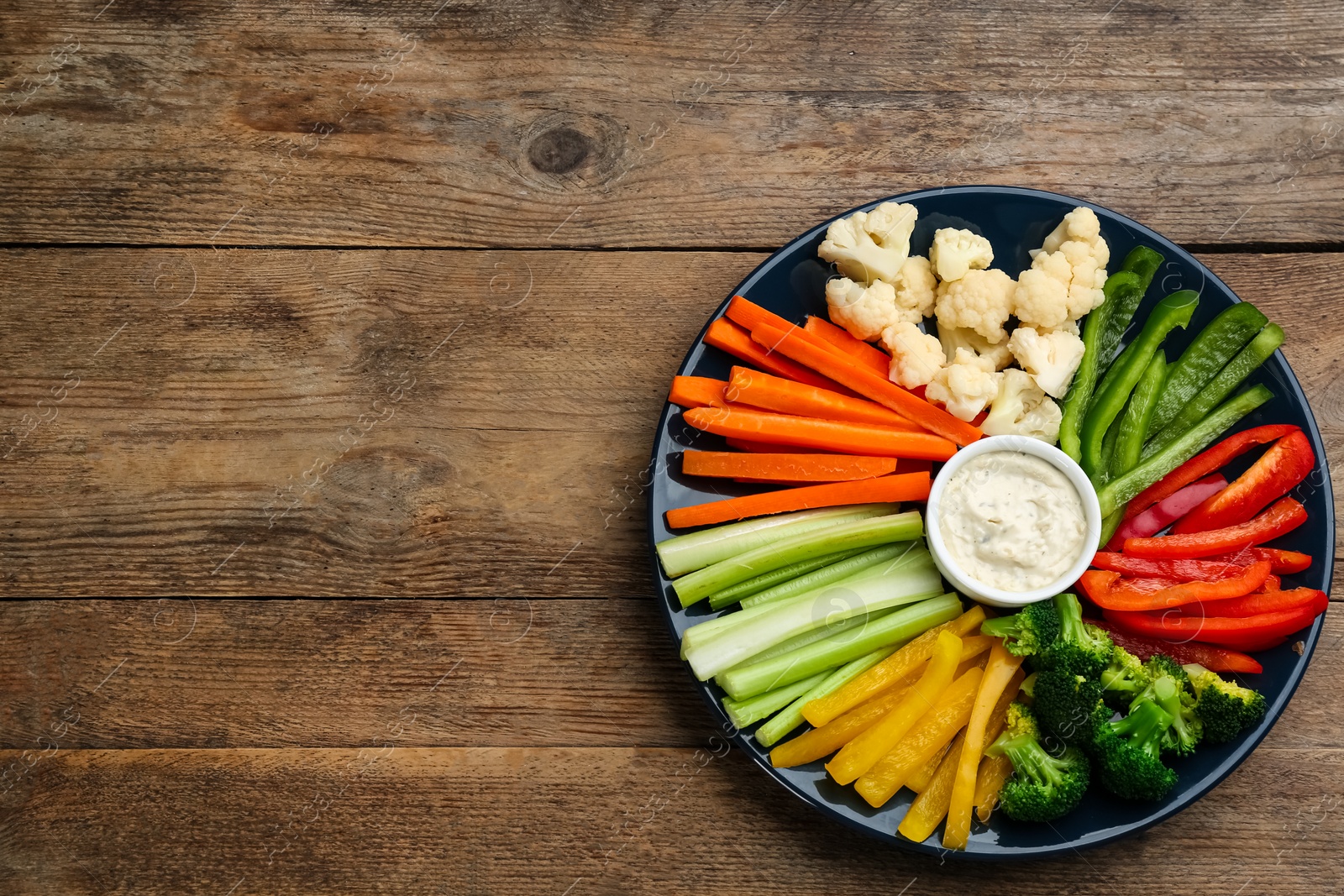 Photo of Plate with celery sticks, other vegetables and dip sauce on wooden table, top view. Space for text