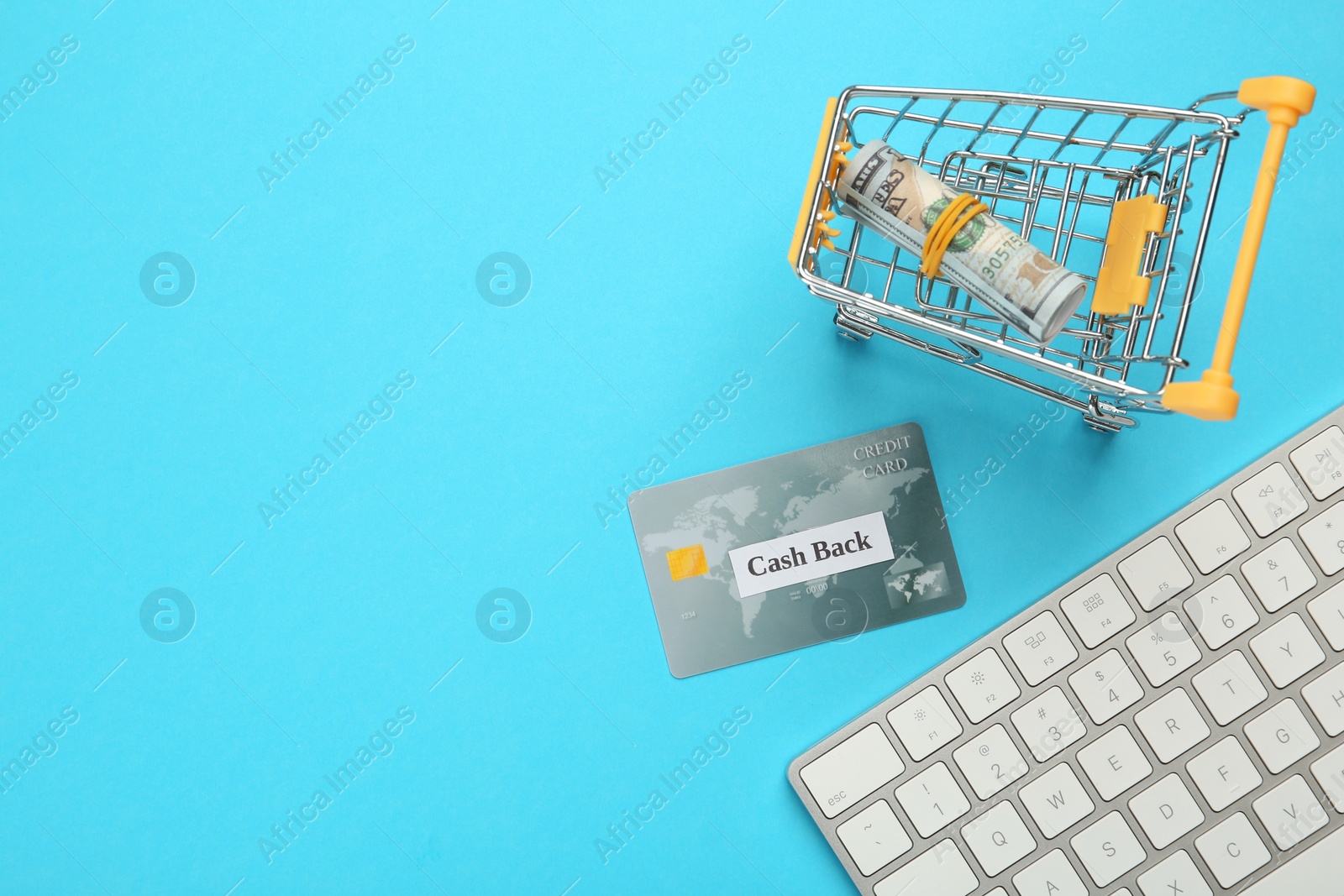 Photo of Keyboard, credit card and rolled dollar banknotes in shopping cart on light blue background, flat lay with space for text. Cashback concept