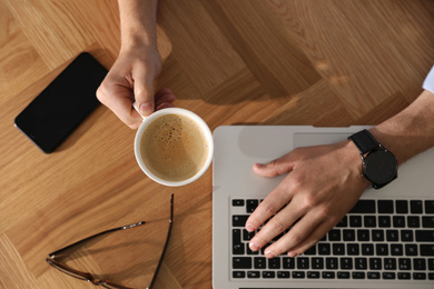 Man with laptop, cup of coffee and smartphone at wooden table, top view