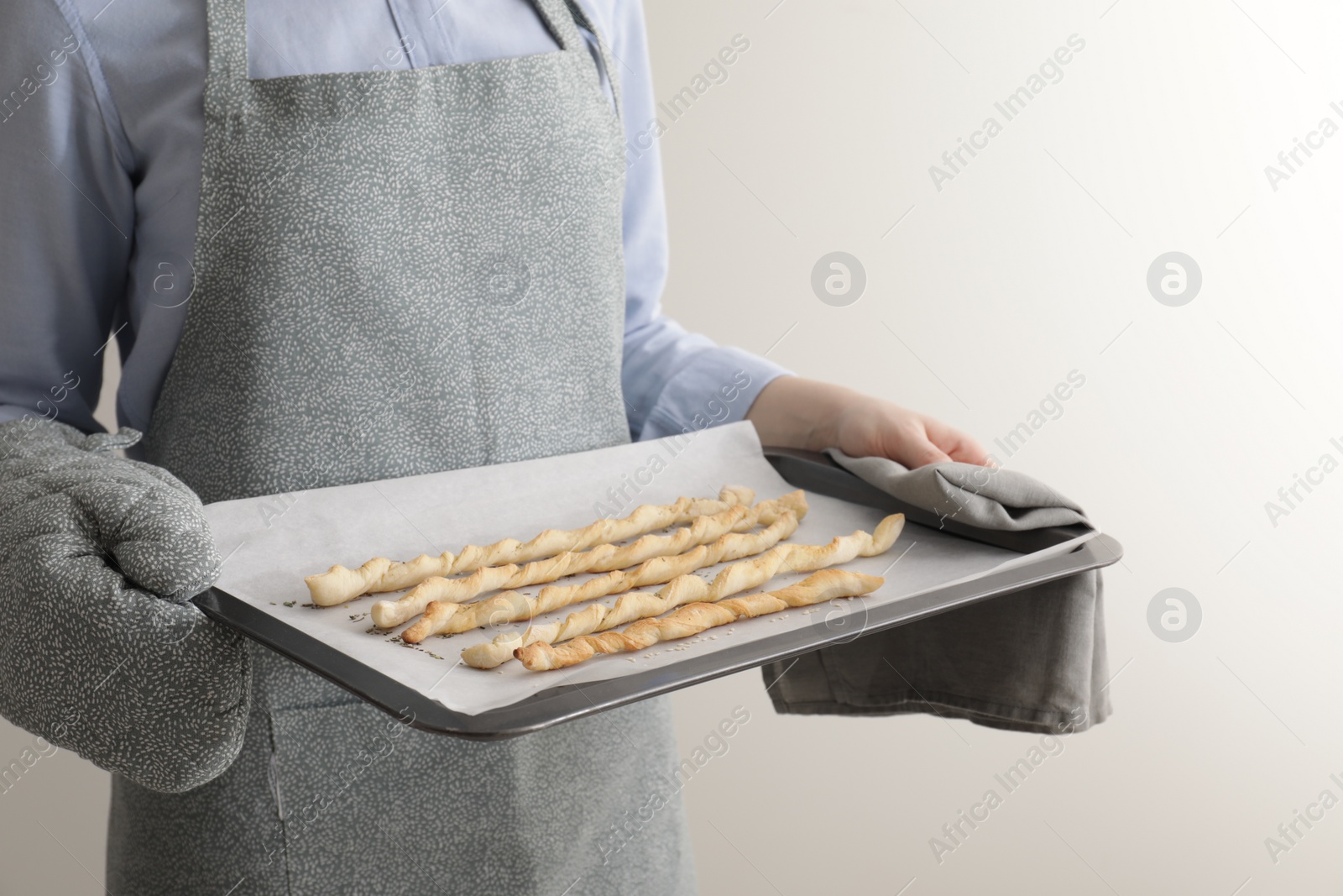 Photo of Woman holding baking sheet with homemade breadsticks on light grey background, closeup. Cooking traditional grissini