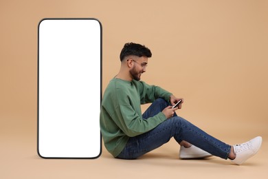 Image of Man with mobile phone sitting near huge device with empty screen on dark beige background. Mockup for design