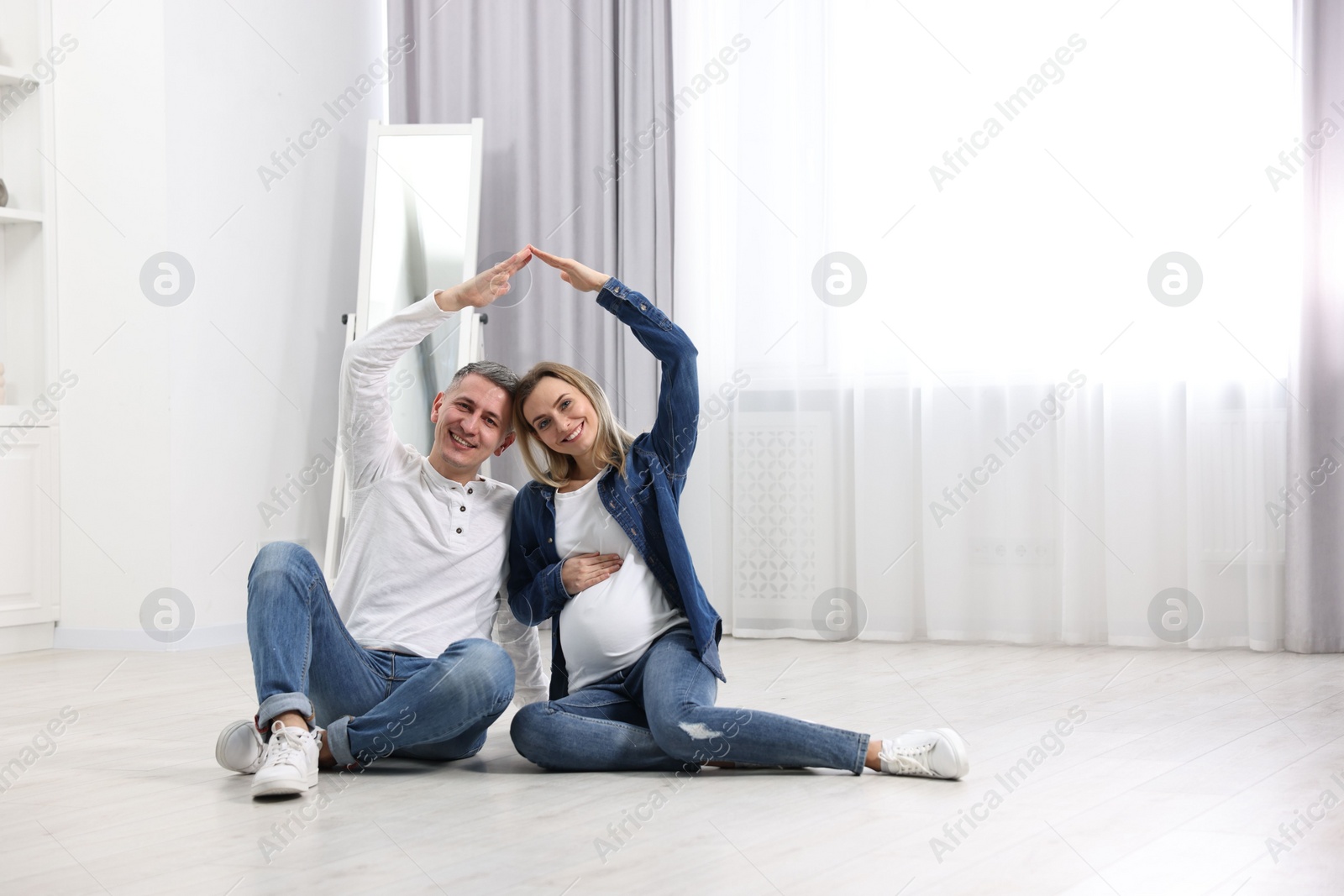 Photo of Pregnant woman with her husband forming roof with their hands while sitting on floor indoors. Space for text